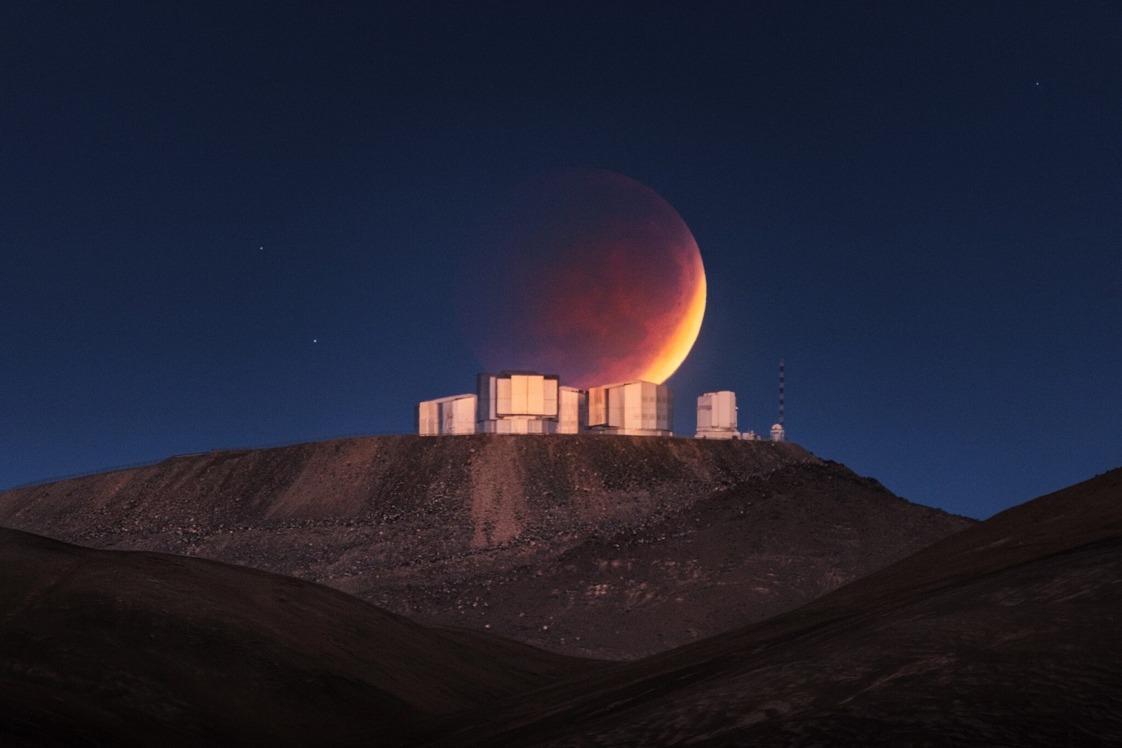 Like Atlas holding up the sky in the Ancient Greek myth, it looks like ESO’s Very Large Telescope (VLT) is supporting the Moon in this extraordinary picture of a total lunar eclipse at the Paranal Observatory in Chile’s Atacama Desert. The photograph was taken on 26 May 2021 by astrophotographer Yuri Beletsky and it is featured in the ESO Calendar 2022. To take this Picture of the Week, some detailed planning was required on the part of the photographer. Careful calculations allowed him to pinpoint a location nine kilometres to the east of Paranal, where the setting lunar eclipse would be briefly visible just above the domes of the telescopes, glowing faintly in the early morning twilight. A total lunar eclipse occurs when the Sun, Earth and Moon align, with the Earth between the Sun and Moon. The Earth’s shadow blocks direct sunlight, so that the only light reflected from the Moon’s surface has been refracted by Earth’s atmosphere, giving the Moon a reddish colour. Since sunlight is filtered by Earth’s atmosphere, astronomers often use lunar eclipses to understand the signatures that life can imprint on the atmosphere, which is extremely useful in our quest for life on other planets. This image is featured in the month of April in the ESO Calendar 2022. If you want to buy a copy, you can find it on our online ESOshop. Deliveries will start in mid September. The calendar measures 49 x 39 cm when packed and has 14 pages, with a cardboard back. It is delivered in a cardboard box. Inside, Lunar phases are also indicated and stunning astronomical images together with breathtaking pictures of ESO&#8217;s telescopes and landscapes will accompany you each month.
